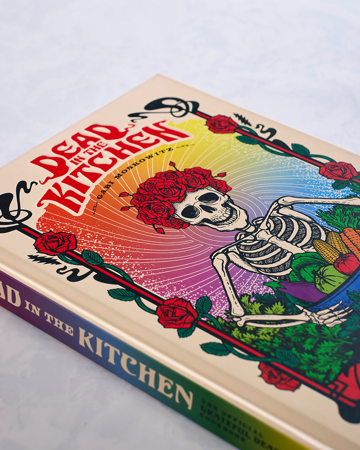 Angled close-up of the “Dead in the Kitchen” cookbook cover, showing the skeleton and rose artwork with gold title lettering on the spine.