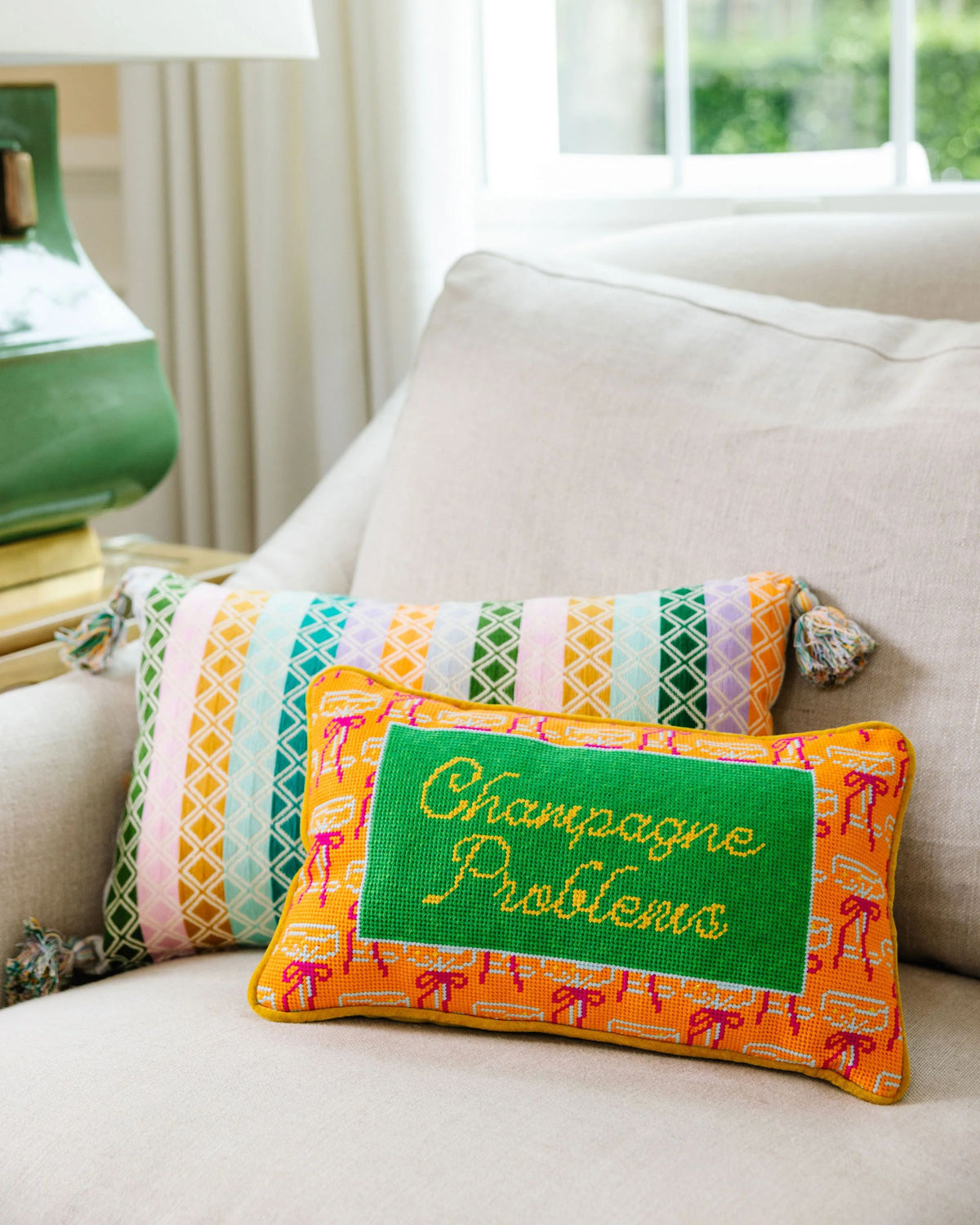 Rectangular embroidered pillow reading “Champagne Problems” on a sofa, with a multicolored geometric pillow behind it.