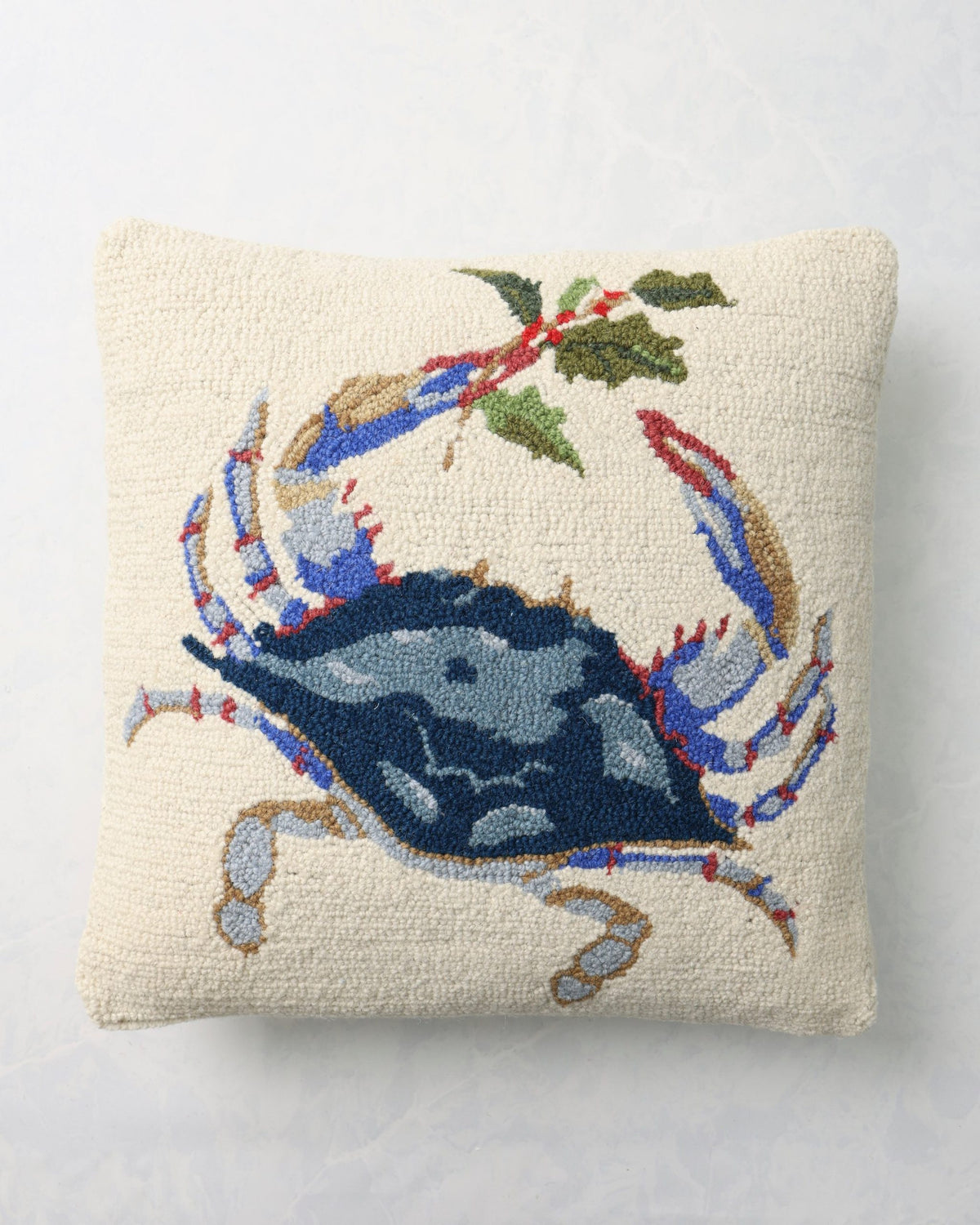 Front of a square hooked pillow with a blue crab, greenery, and red accents.