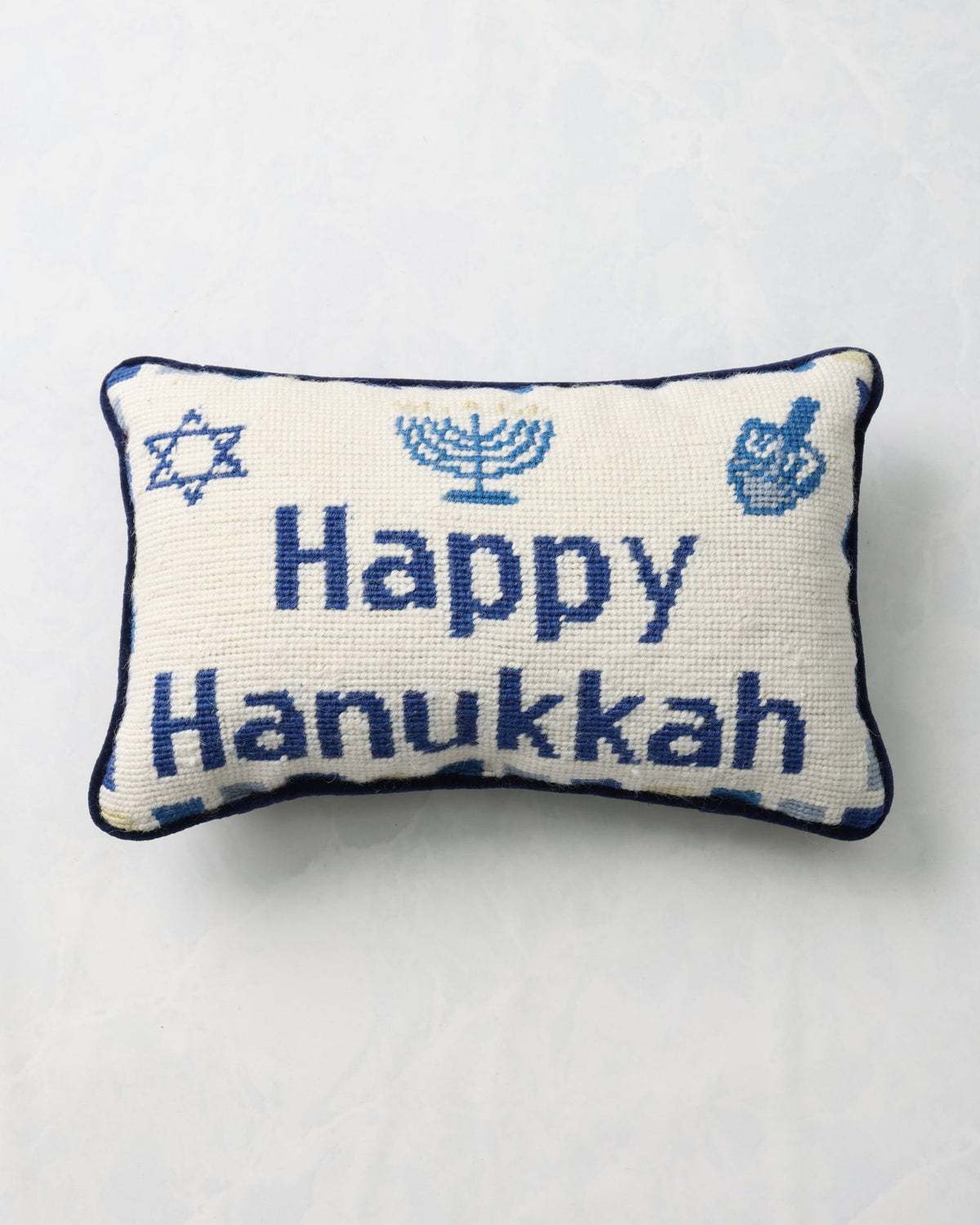 Front of an ivory needlepoint pillow reading “Happy Hanukkah,” with a Star of David, menorah, and dreidel motifs.