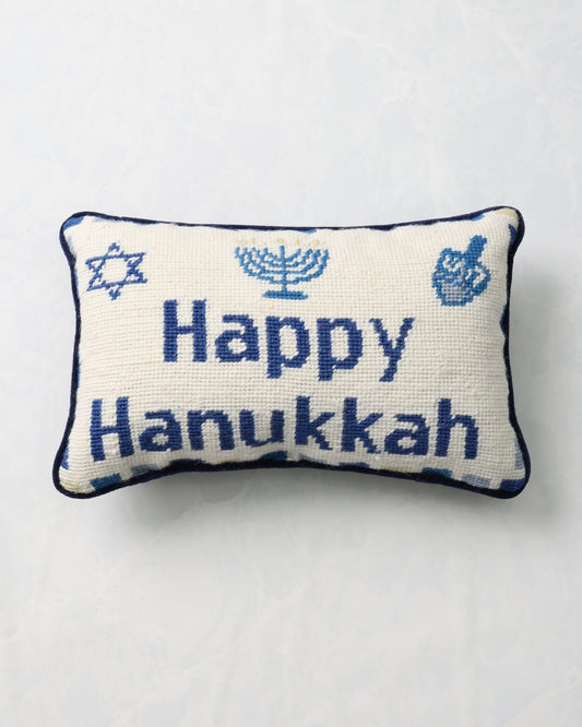 Front of an ivory needlepoint pillow reading “Happy Hanukkah,” with a Star of David, menorah, and dreidel motifs.