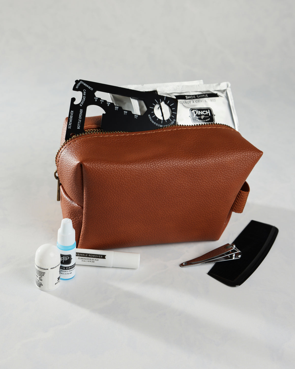 Open cognac Hemergency Kit pouch with grooming and repair items displayed around it, including a comb, nail clipper, bottles, and shoe-shine packet.