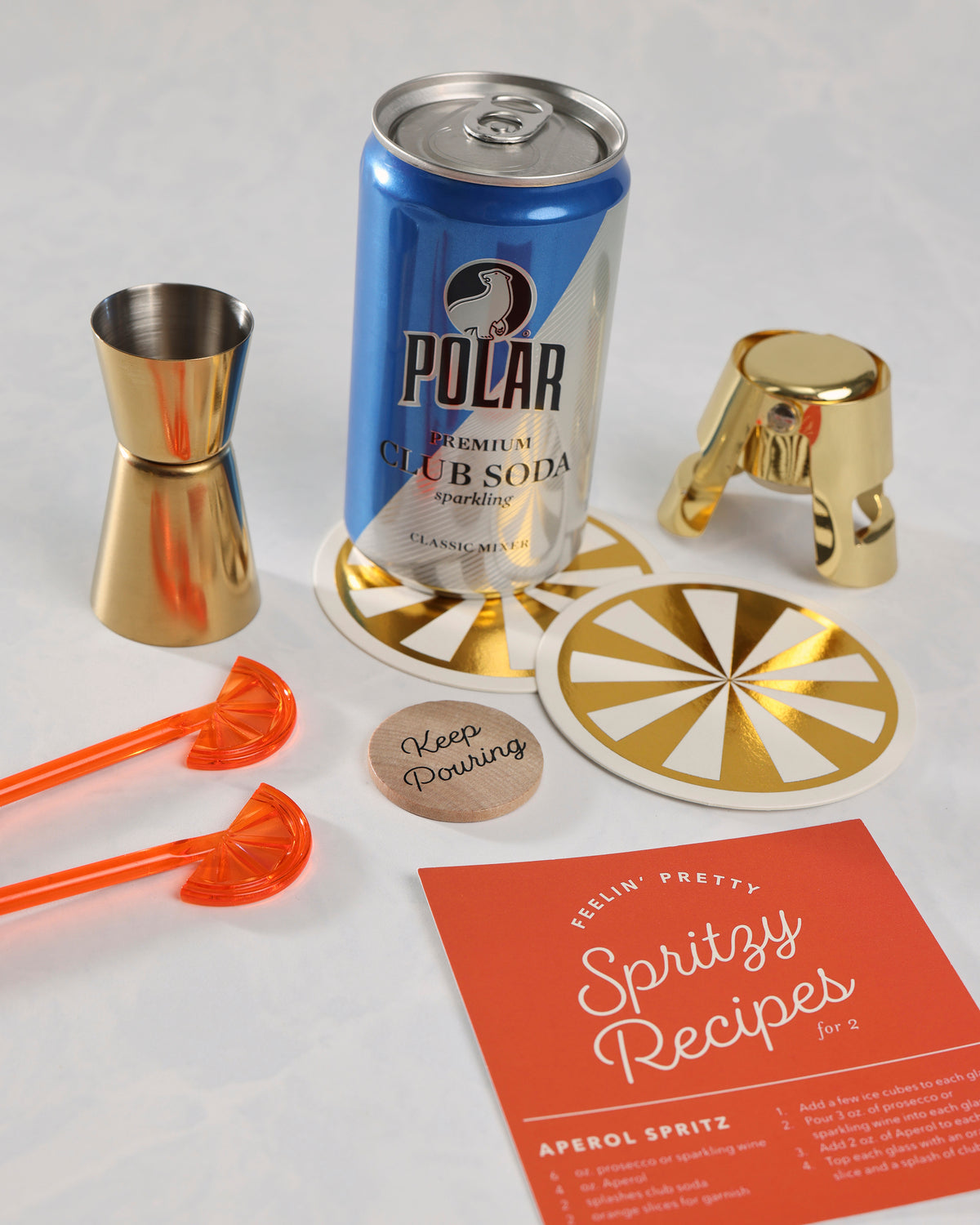 Spritz Kit contents arranged together: Polar club soda, gold jigger, gold bottle stopper, two orange swizzle sticks, two coasters, a decision coin, and a spritz recipe card.