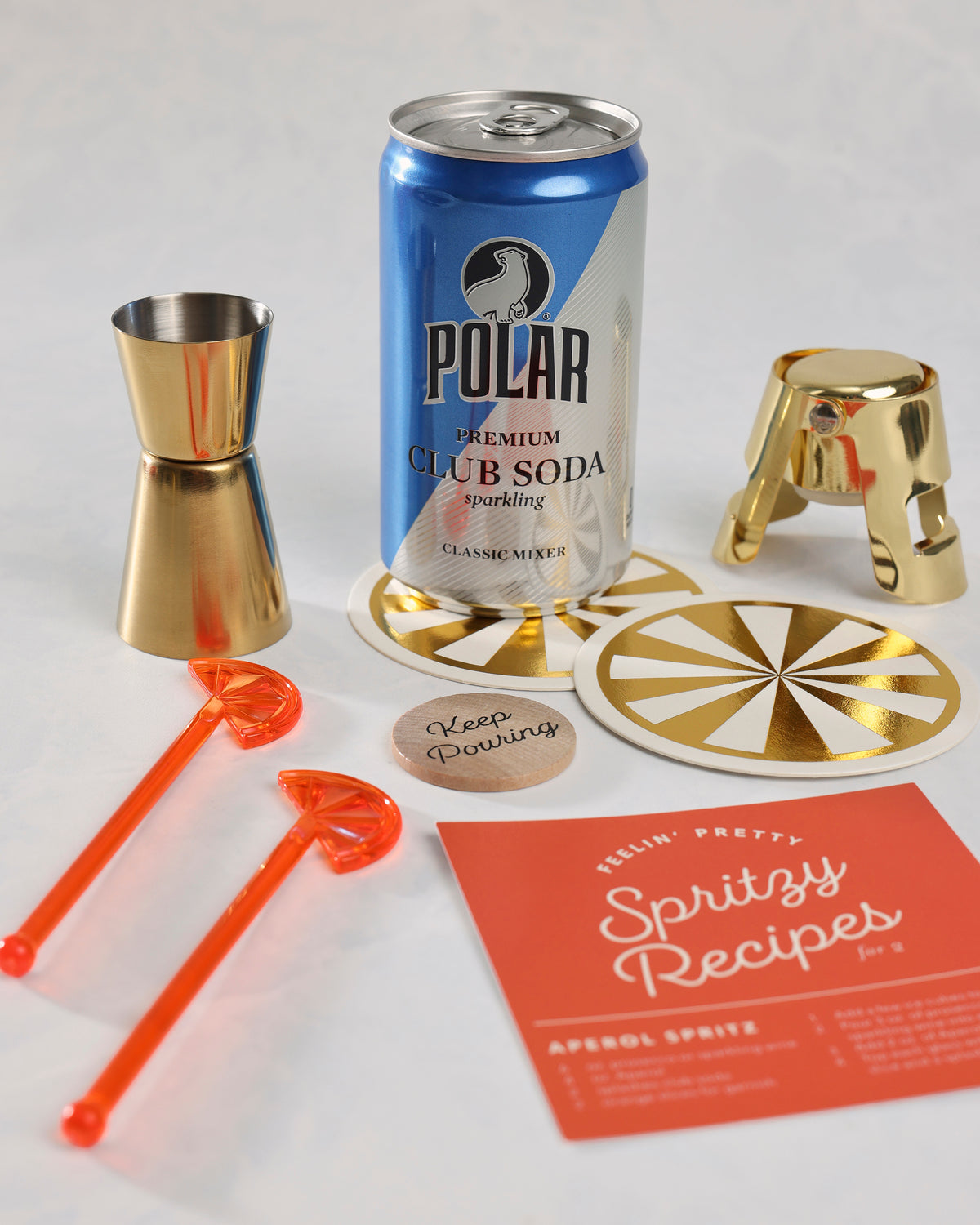 Spritz Kit contents arranged on a surface: Polar club soda, gold jigger, gold bottle stopper, two orange swizzle sticks, two coasters, a decision coin, and a spritz recipe card.