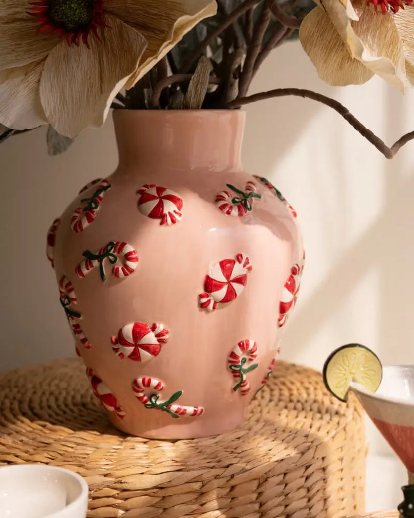Pink ceramic vase with red-and-white peppermint decorations holding dried flowers on a woven tabletop