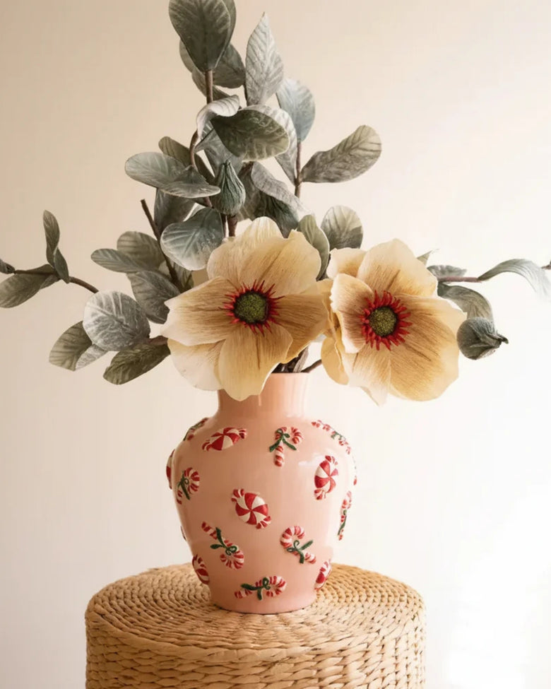 Pink ceramic vase decorated with peppermint candies and candy canes, holding two pale yellow flowers and leafy stems on a woven stand.