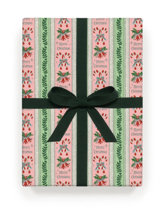 Ribbon-wrapped roll of pink Christmas wrapping paper with vertical green and cream stripes, candy canes, bells, holly, and repeated “Merry Christmas” text.