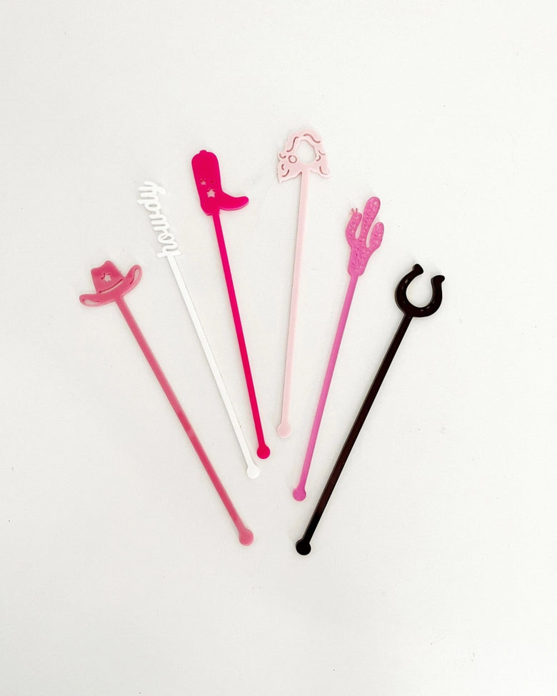 Six colorful Western-themed stir sticks arranged on a white background, including cowboy hats, a boot, a cactus, a horseshoe, and a white script-shaped design.