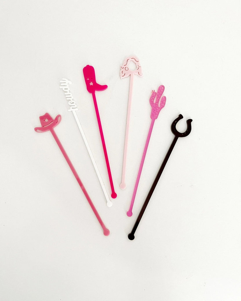 Six colorful Western-themed stir sticks arranged on a white background, including cowboy hats, a boot, a cactus, a horseshoe, and a white script-shaped design.