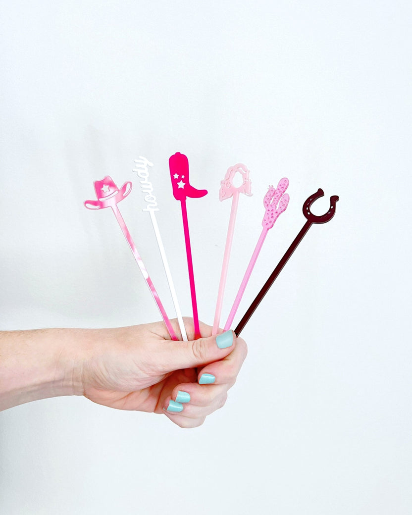 A hand holds six pink, white, and dark red western-themed acrylic stir sticks, including cowboy hat, boot, and horseshoe shapes.