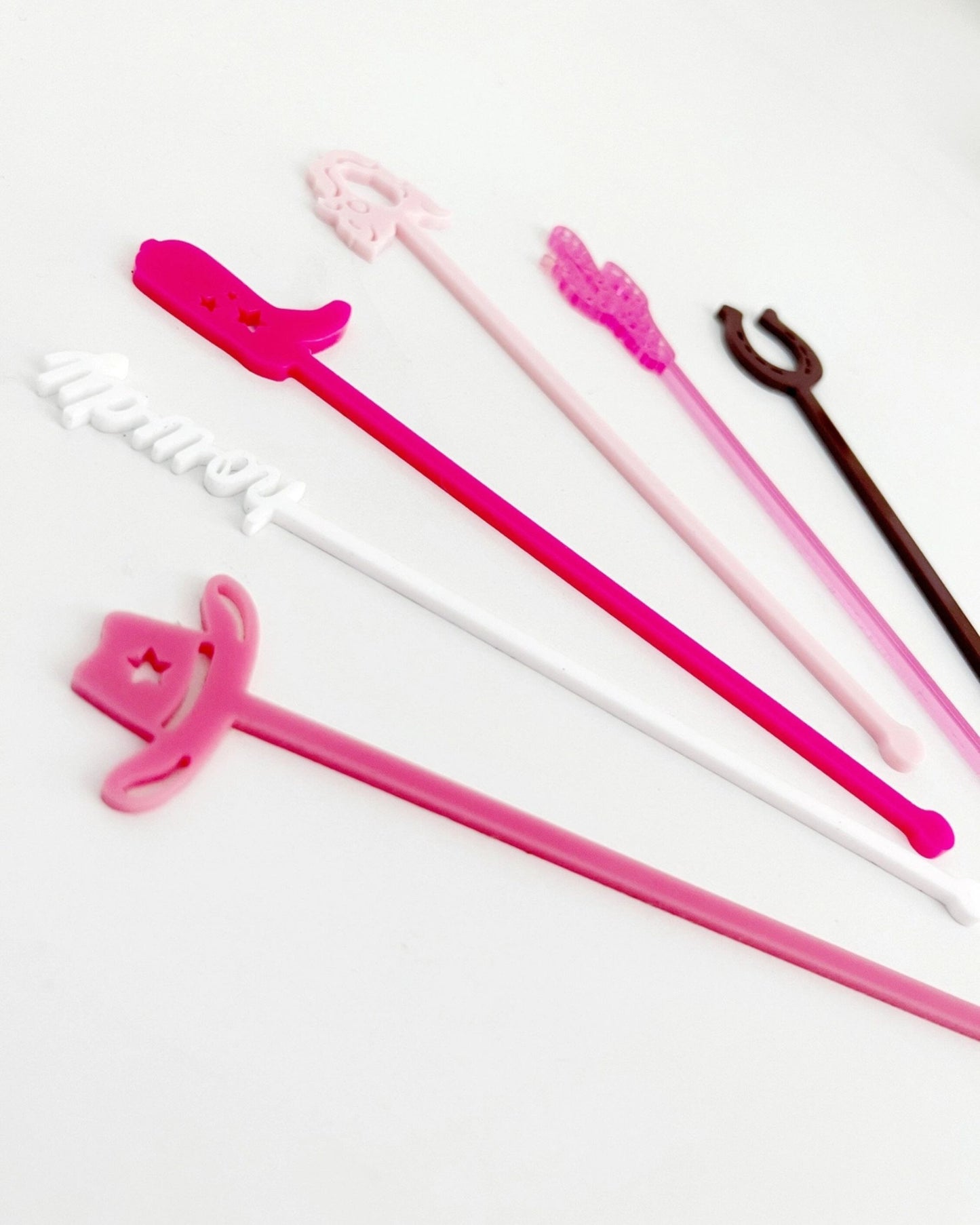 Close-up of six colorful Western-themed stir sticks arranged diagonally on a white background, showing cowboy hats, a boot, a cactus, a horseshoe, and a white script-shaped design.