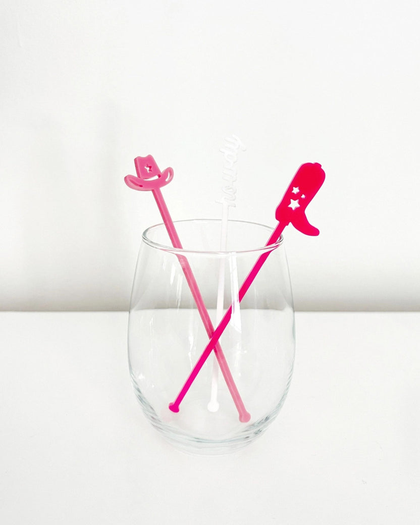 Three pink and white Western-themed cocktail stir sticks crossed inside a clear drinking glass.