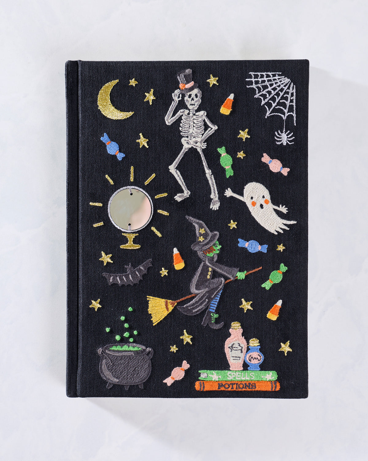 Front cover of a black embroidered journal decorated with Halloween motifs including a skeleton, ghost, witch, moon, stars, candies, spiderweb, and cauldron.