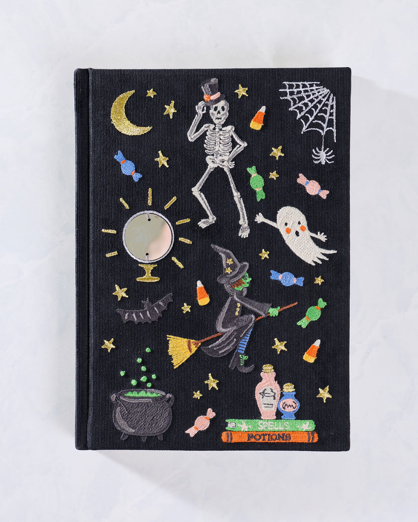 Front cover of a black embroidered journal decorated with Halloween motifs including a skeleton, ghost, witch, moon, stars, candies, spiderweb, and cauldron.