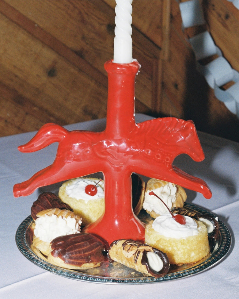 Red horse-shaped candlestick holder supporting a white taper candle, surrounded by pastries on a serving plate.