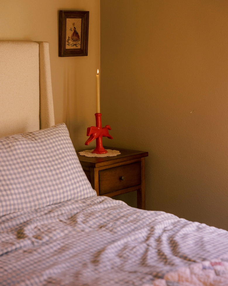 Red horse-shaped candlestick holder with a lit taper candle on a bedside table beside a bed.