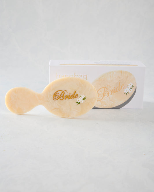 Cream handbag hairbrush with gold “Bride” lettering and small white flower decoration, displayed with its packaging.