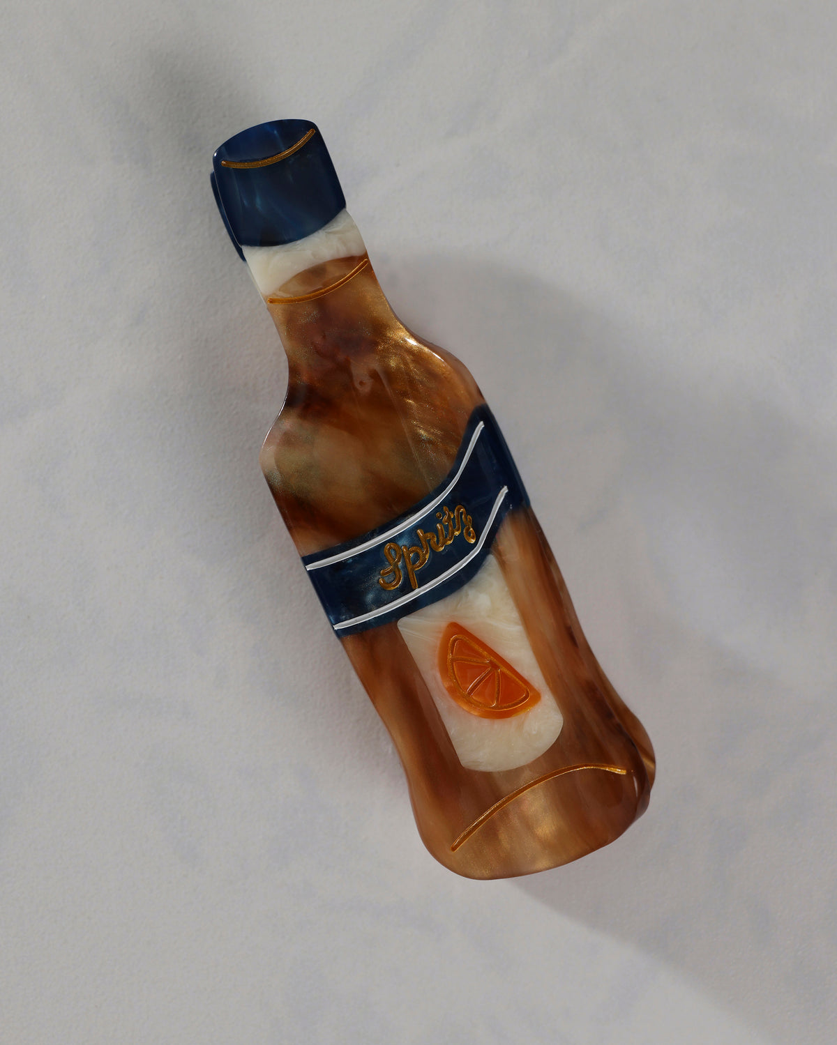 Front view of a hand-painted citrus spritz claw hair clip shaped like a bottle, with navy, amber, and cream sections, a citrus emblem, and the word “Spritz.”