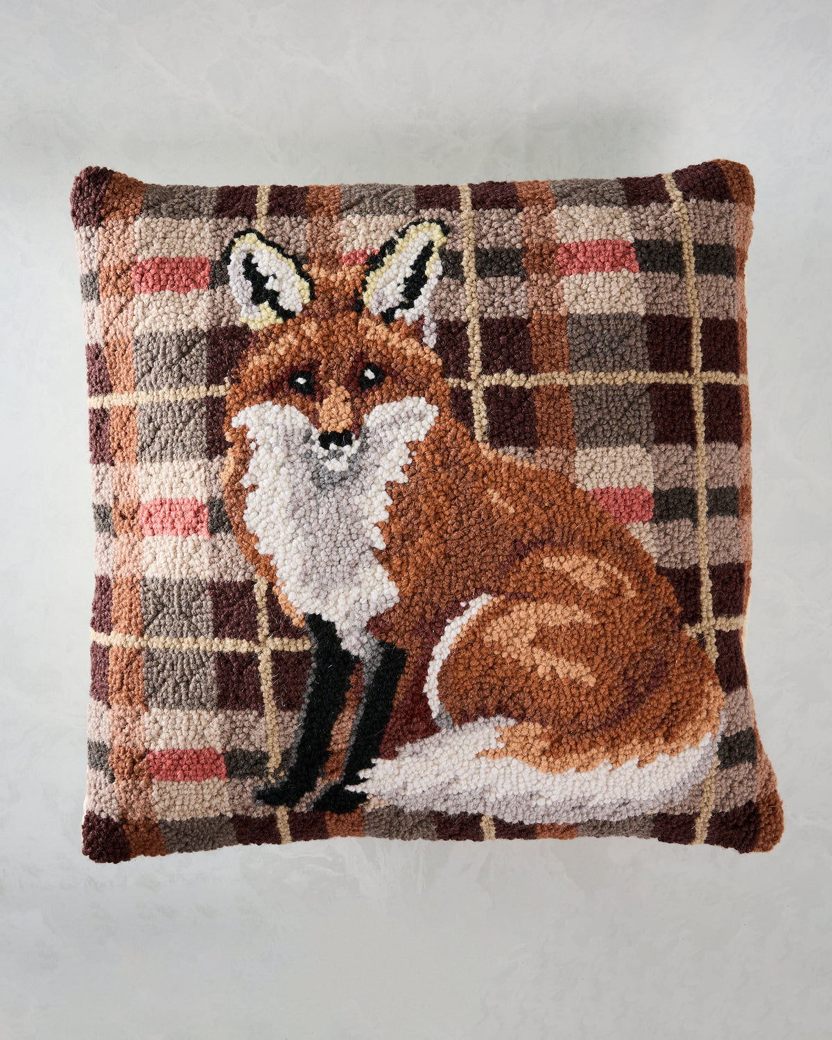 Front of a square hooked pillow featuring a seated fox on a plaid background.