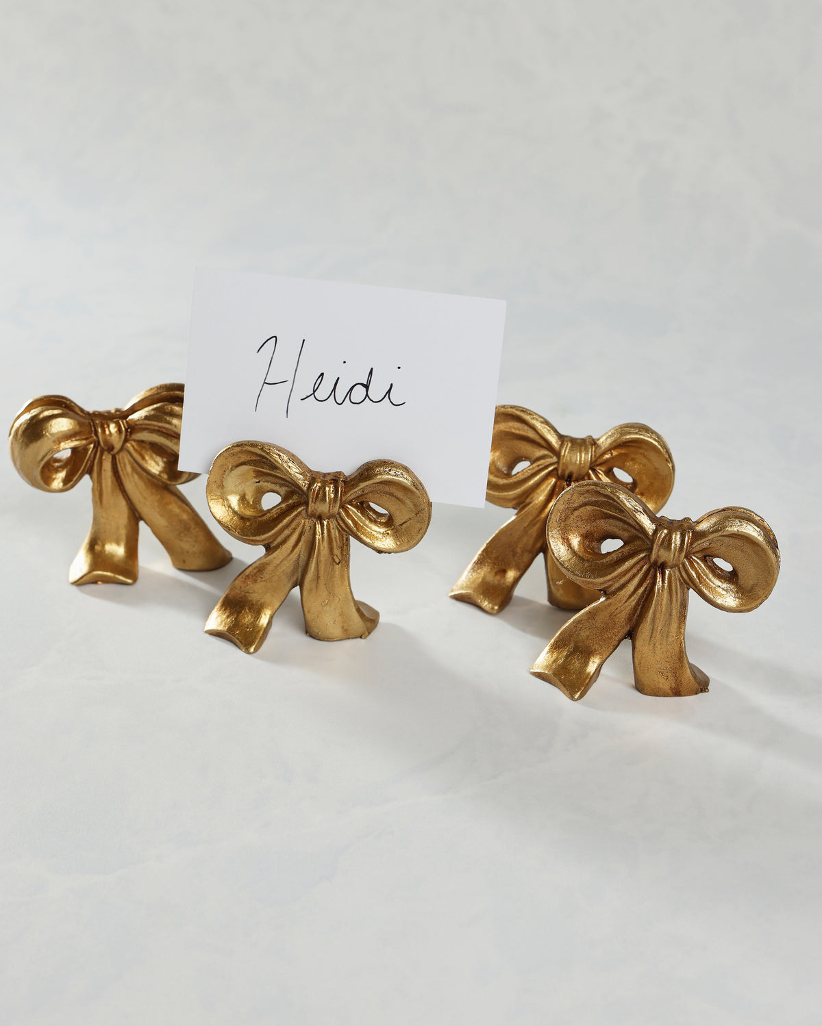 Four gold bow placecard holders, with one partly covered by a card labeled “Heidi.”