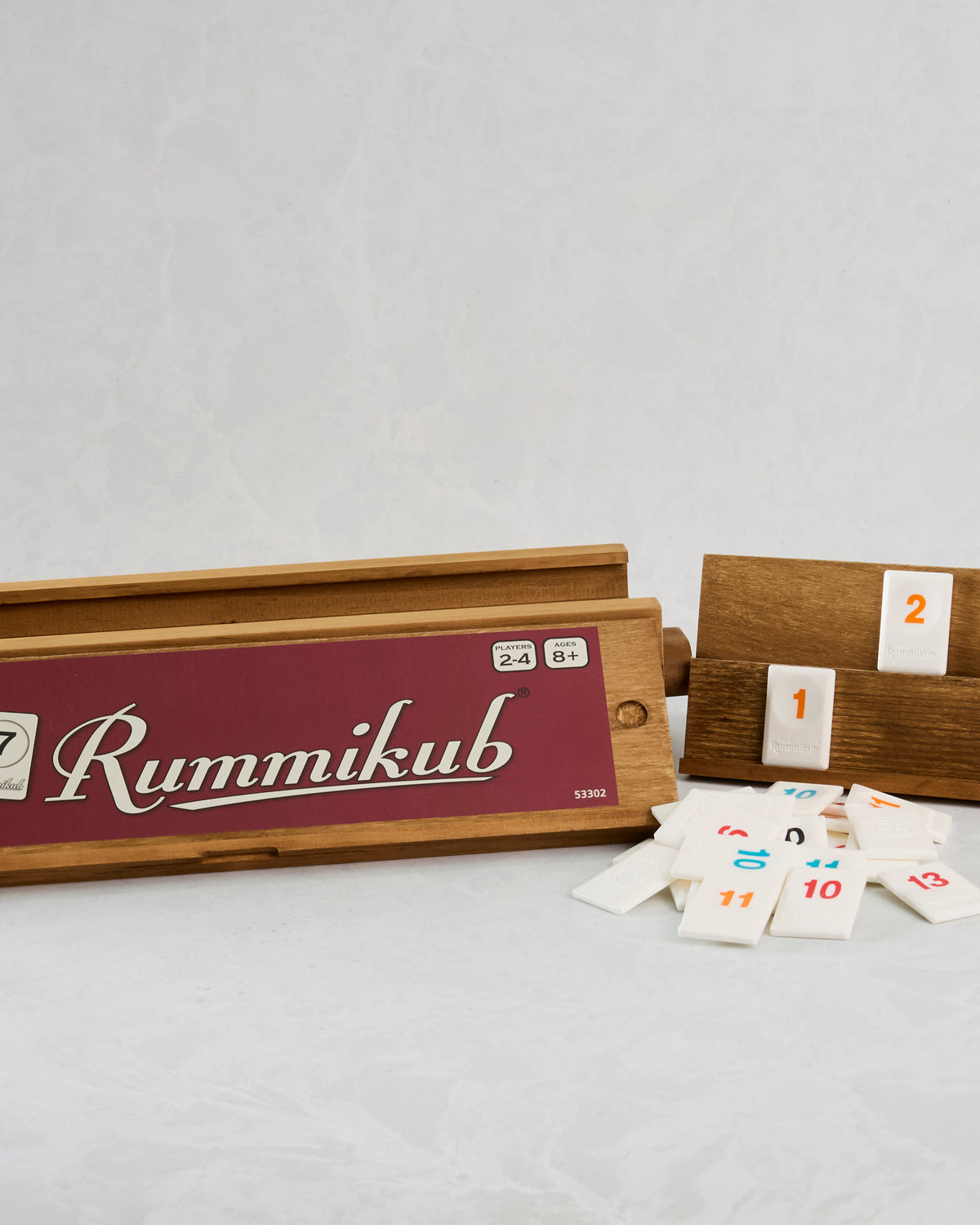 Open wooden Rummikub game box with tile racks and scattered numbered tiles.