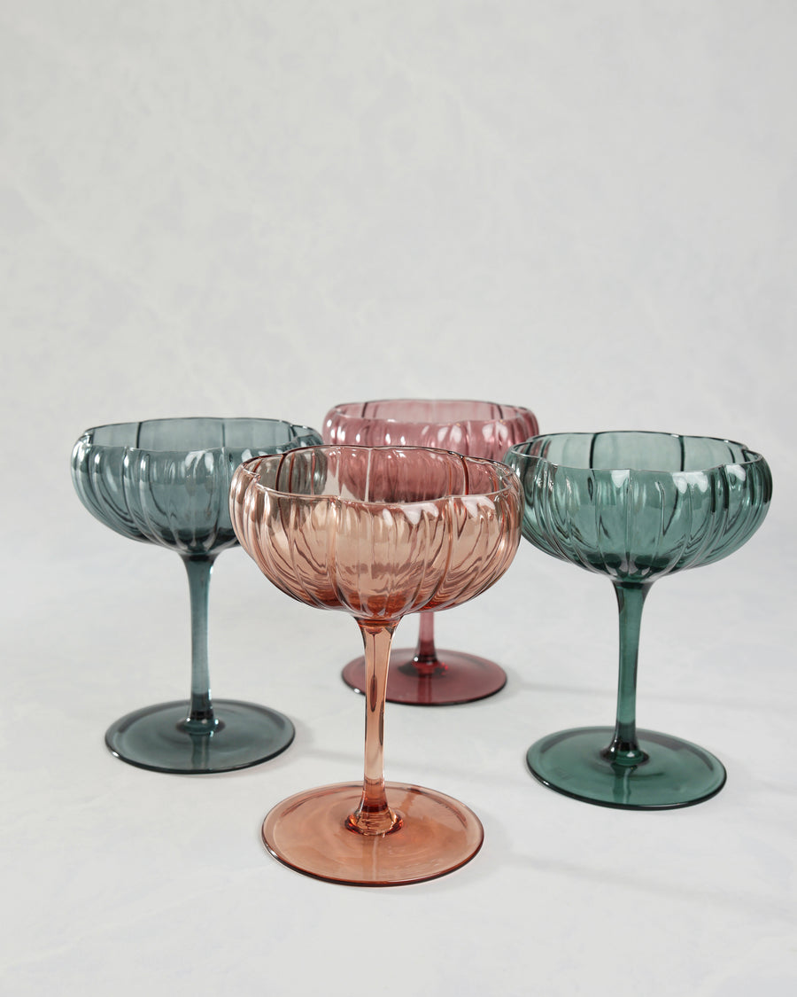 Four flower-shaped coupe glasses in pink, peach, blue, and green tones shown from a lower angle.