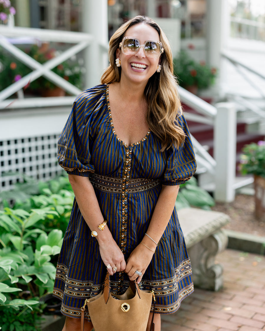 Person wearing a blue and olive striped above-the-knee dress and holding a tan handbag outdoors.