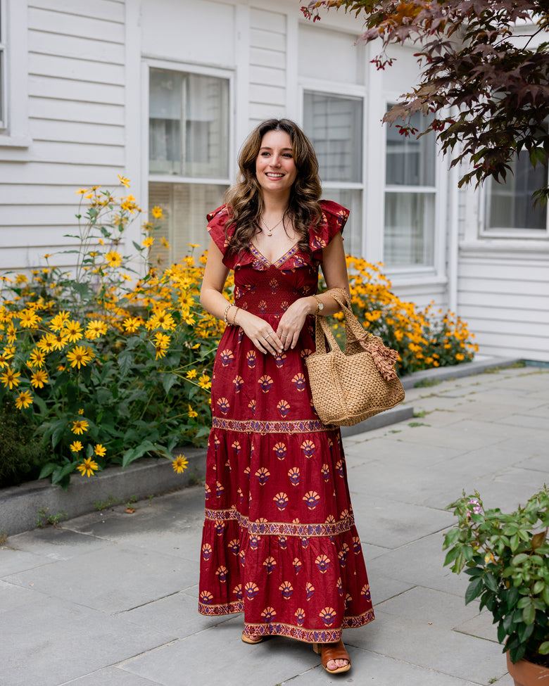 Person wearing a red patterned maxi dress standing beside yellow flowers and carrying a woven tote.