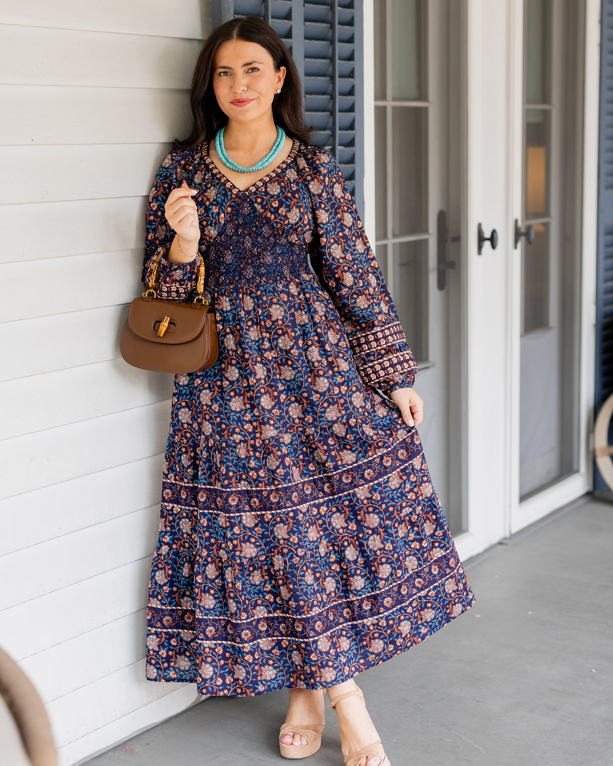Person wearing a navy patterned long-sleeved maxi dress with a brown handbag by a doorway.