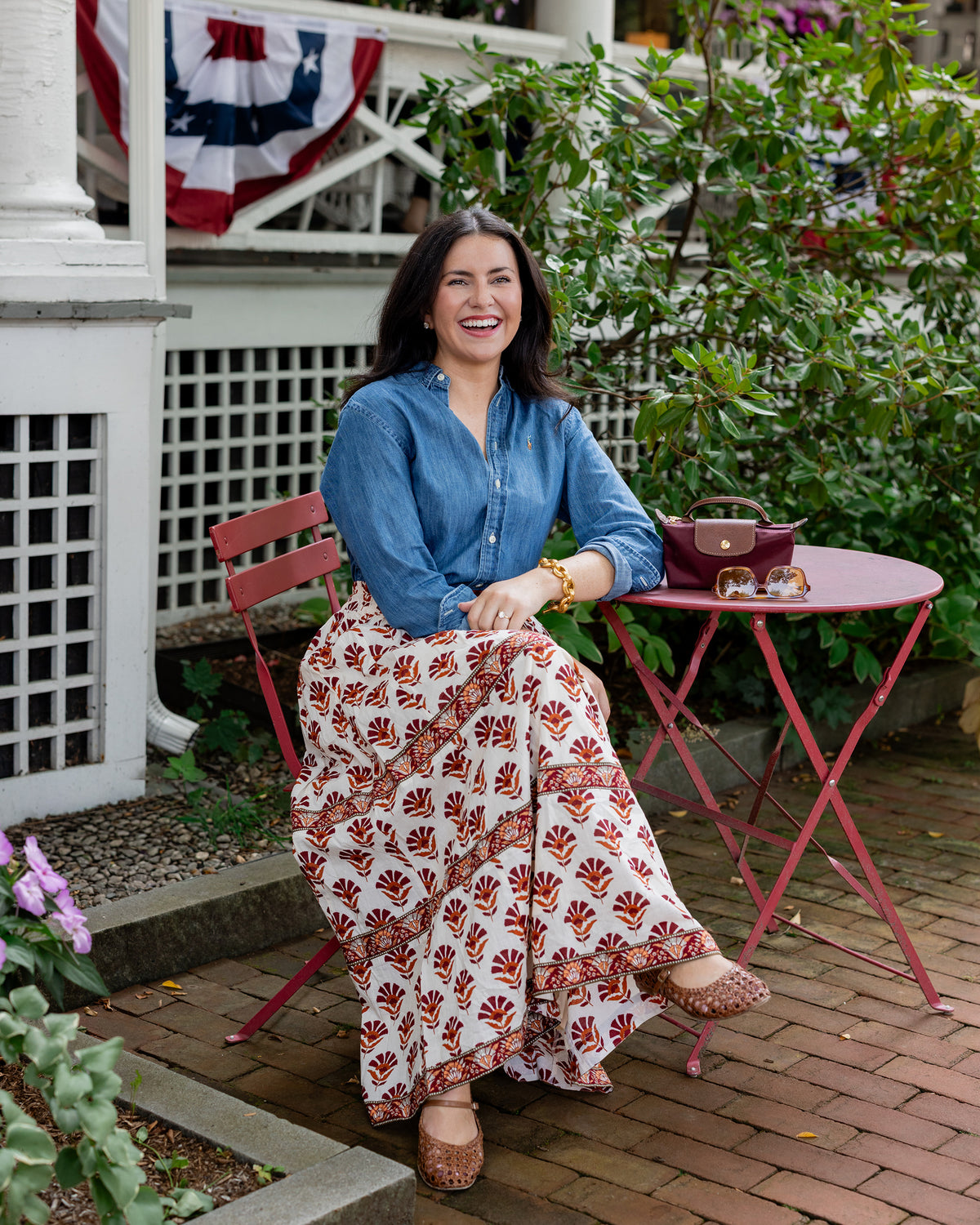 Long cream Sierra Bloom skirt with a red floral print, shown worn with a blue shirt while seated outdoors.