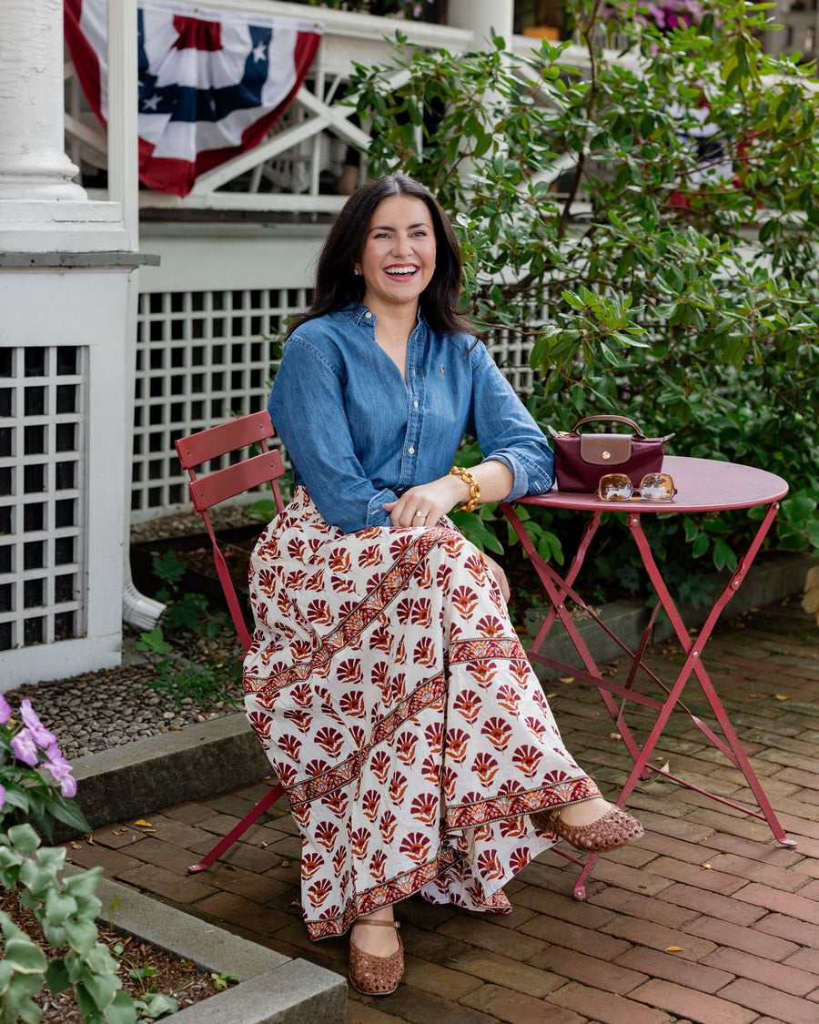 Long cream Sierra Bloom skirt with a red floral print, shown worn with a blue shirt while seated outdoors.