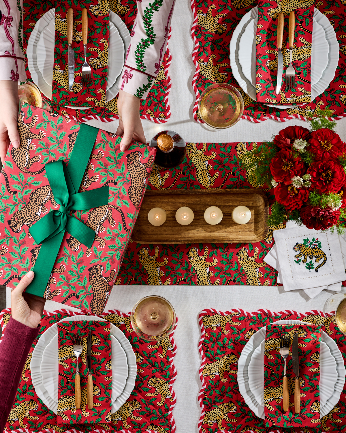 Overhead view of a holiday table set with patterned placemats, plates, cutlery, drinks, candles, flowers, and a wrapped gift.