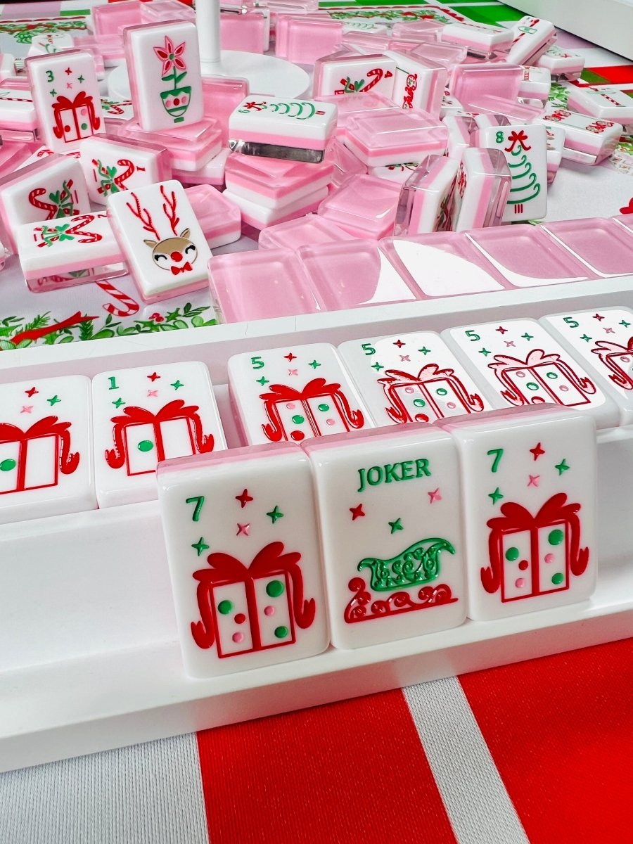 Holiday mahjong tiles arranged in a display rack with pink cases behind them, showing wrapped-gift tiles and a joker tile.