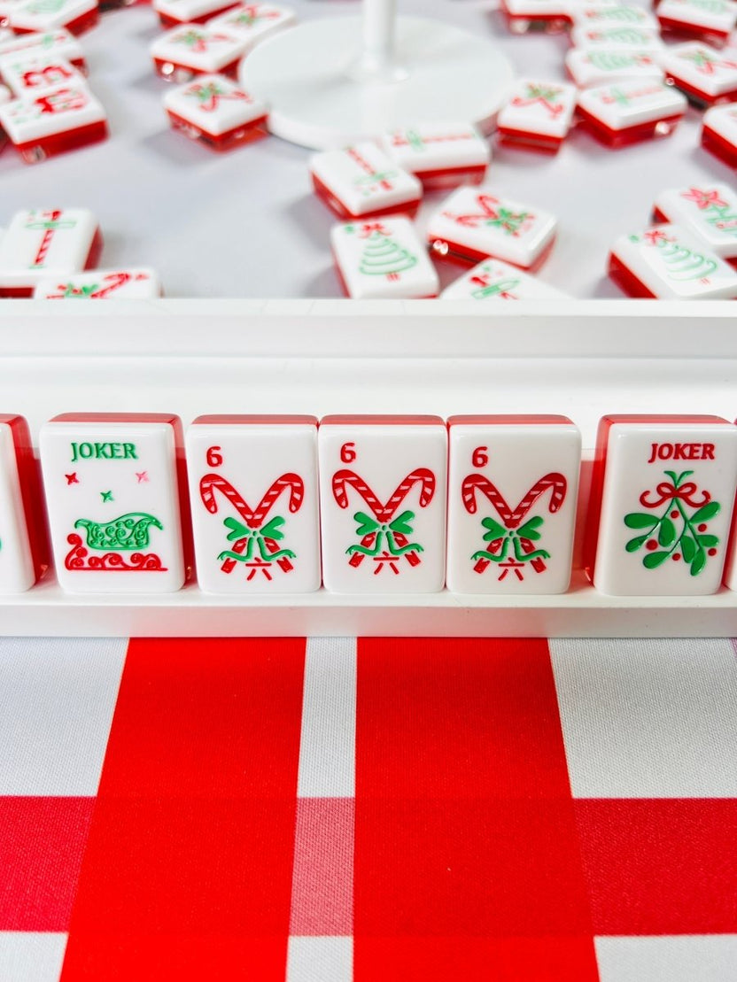 Holiday mahjong tiles arranged around a white circular stand, with a front row displaying joker, candy cane, and mistletoe designs.