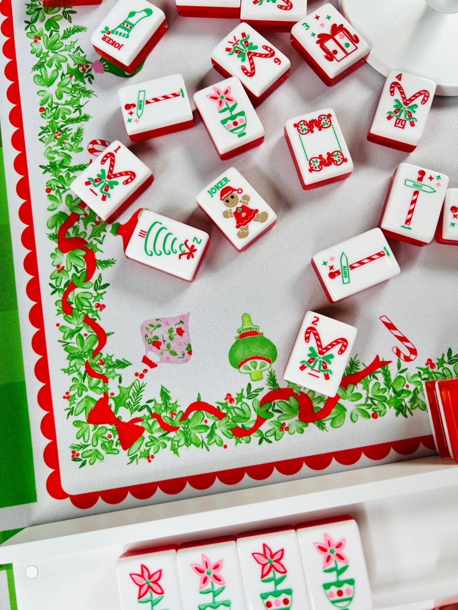Holiday mahjong tiles scattered on a Christmas-themed table covering, showing candy canes, wrapped gifts, Christmas trees, and joker designs.