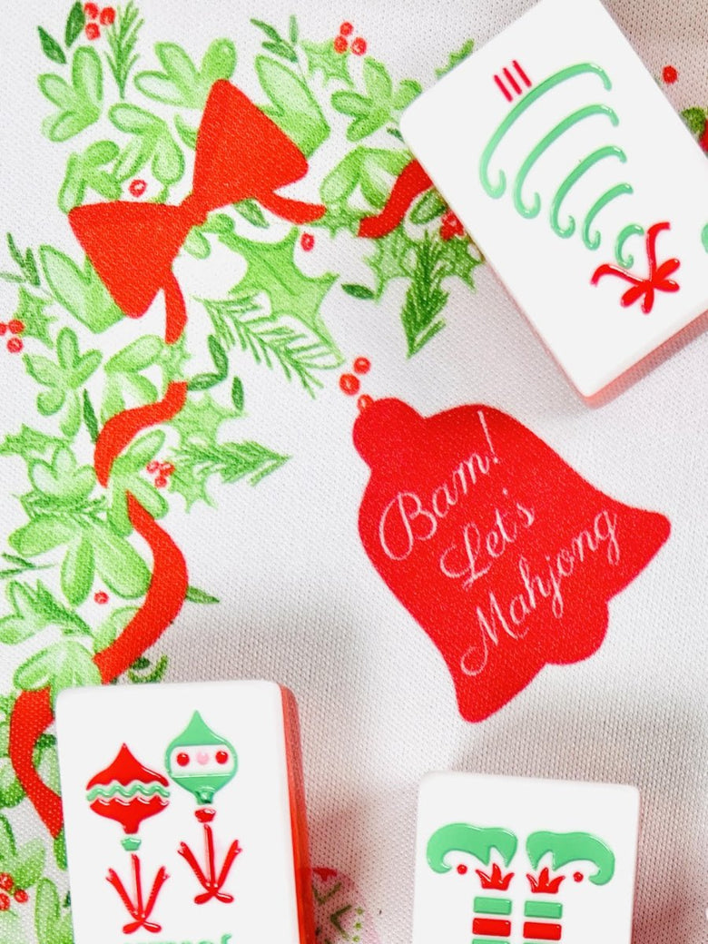 Close-up of the Holly Jolly Christmas Mahjong mat showing a red ribbon, holiday greenery, a tile with green curved symbols, and a red bell reading “Bam! Let’s Mahjong.”