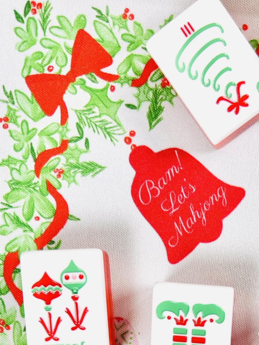 Close-up of the Holly Jolly Christmas Mahjong mat showing a red ribbon, holiday greenery, a tile with green curved symbols, and a red bell reading “Bam! Let’s Mahjong.”